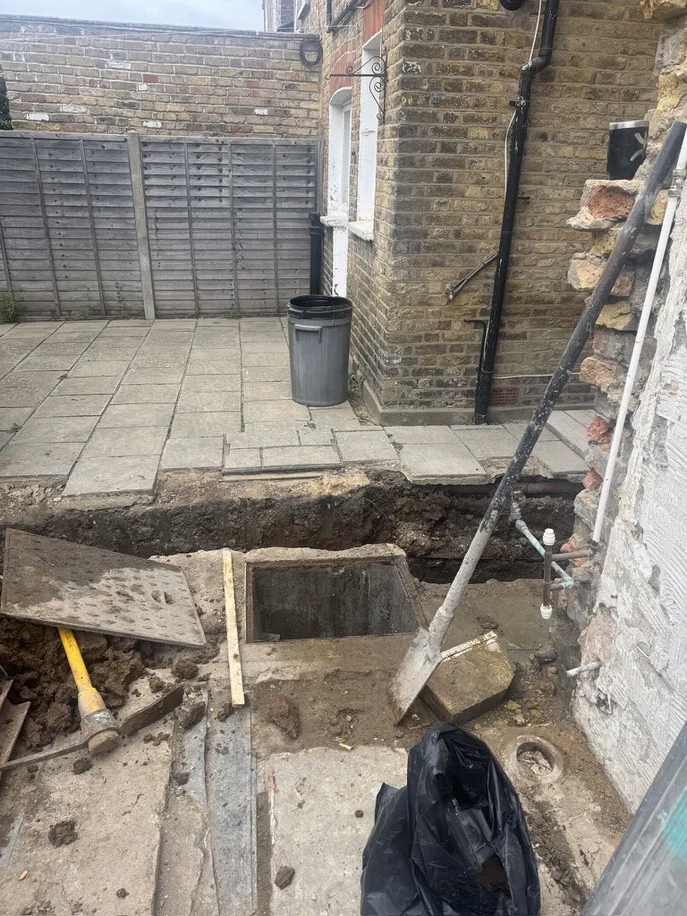 Rear patio excavated to expose the manhole and drain runs before extension foundations, with new pipework at the wall